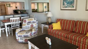 A living room with a striped sofa, a coffee table, a chair with beach-themed upholstery, a dining table with chairs, a kitchen area, and a framed picture on the wall. - Elevated First Floor Beachfront w/ Beautiful Balcony Views (Gulf Shores)