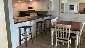A compact kitchen with white cabinets, a central island with stools, and a dining table with chairs. - Elevated First Floor Beachfront w/ Beautiful Balcony Views (Gulf Shores)