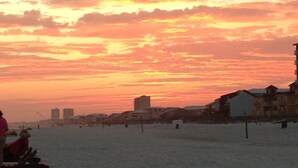 A beach scene with a sunset, buildings in the background, and wooden structures on the sand. - Elevated First Floor Beachfront w/ Beautiful Balcony Views (Gulf Shores)