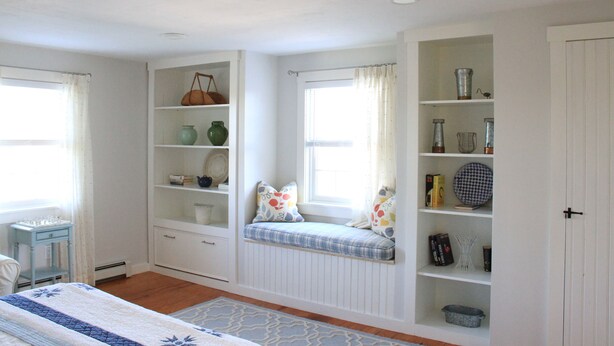 Une chambre à coucher comprenant un lit, un coin fenêtre avec des coussins, une bibliothèque intégrée et une armoire.