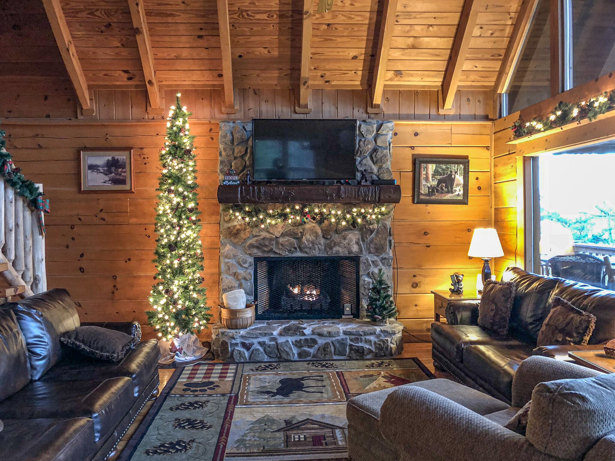 L’intérieur chaleureux d’un chalet avec une cheminée en pierre, des canapés en cuir, un sapin de Noël et un téléviseur fixé au mur.