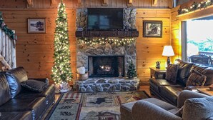 Ein gemütliches Blockhaus-Interieur mit Kamin aus Stein, Ledersofas, einem Weihnachtsbaum und einem an der Wand befestigten Fernseher.