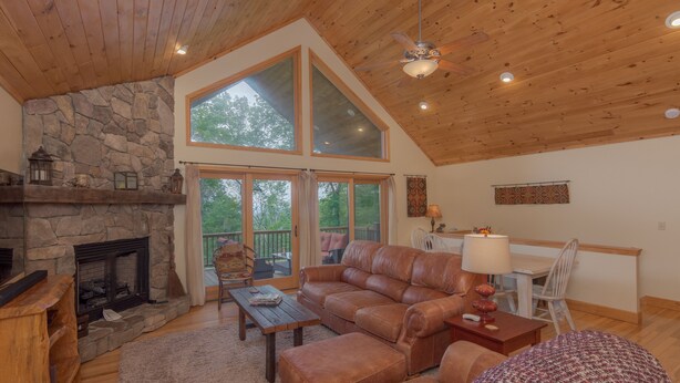 A spacious living room with a stone fireplace, leather sofa, and a large window overlooking a deck. - Get-Away Mountain Cabin. Hot Tub, Sunset Mtn. Views, Game Room, Near Asheville (Mars Hill)