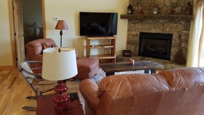 A cozy living room with a stone fireplace, leather furniture, a flat-screen TV, and a wooden ceiling. - Get-Away Mountain Cabin. Hot Tub, Sunset Mtn. Views, Game Room, Near Asheville (Mars Hill)