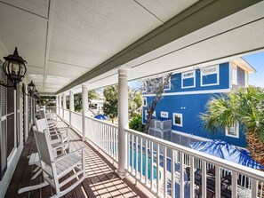 A covered porch with white railings and rocking chairs, overlooking a blue building with a pool.