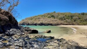 A rocky shoreline with clear turquoise water, a sandy beach, and a forested hill in the background.
