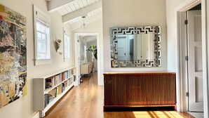 Ein Flur mit Holzboden, einem Bücherregal, einem Spiegel mit geometrischem Rahmen und einer Tür rechts.