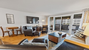 A modern living room with a sofa, armchairs, a coffee table, and a TV. There is a dining table with chairs and a bookshelf. The room has large windows with curtains. - Nye Beach, Restaurants, Shop! Walk to the Beach! (Newport)