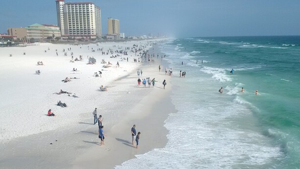 A beach with people walking and swimming, and buildings in the background.