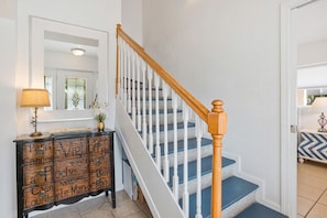 A staircase with white railings and blue steps, a wooden console table with decorative items, and a mirror above the console. - Ultimate Family Home!   Game Porch / No Stairs / Pets / Pool / Tesla Charger (Pensacola Beach)