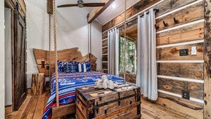 A rustic cabin-style bedroom with a wooden bed, a hanging rope swing, and a large window with curtains.