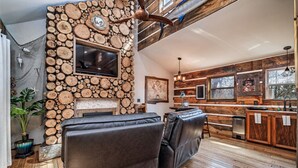A cozy living room with a stone wall, leather sofas, a fireplace, and a kitchen area with wooden cabinets.