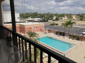 A view of a swimming pool from a balcony with a mesh screen, surrounded by greenery and buildings.