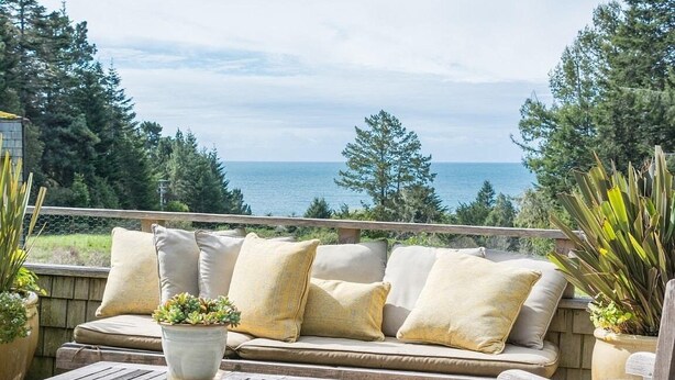 A wooden deck with a beige sofa and cushions, a small table, and potted plants. - Come take in the ocean, gaze at the starry skies, commune with nature. (The Sea Ranch)