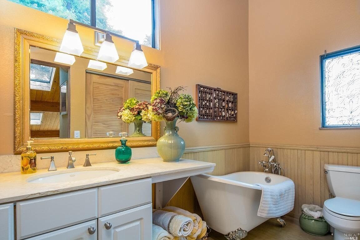 A bathroom with a vanity, a bathtub, and a toilet. There is a mirror with lights above it, a vase with flowers on the vanity, and towels on the floor.