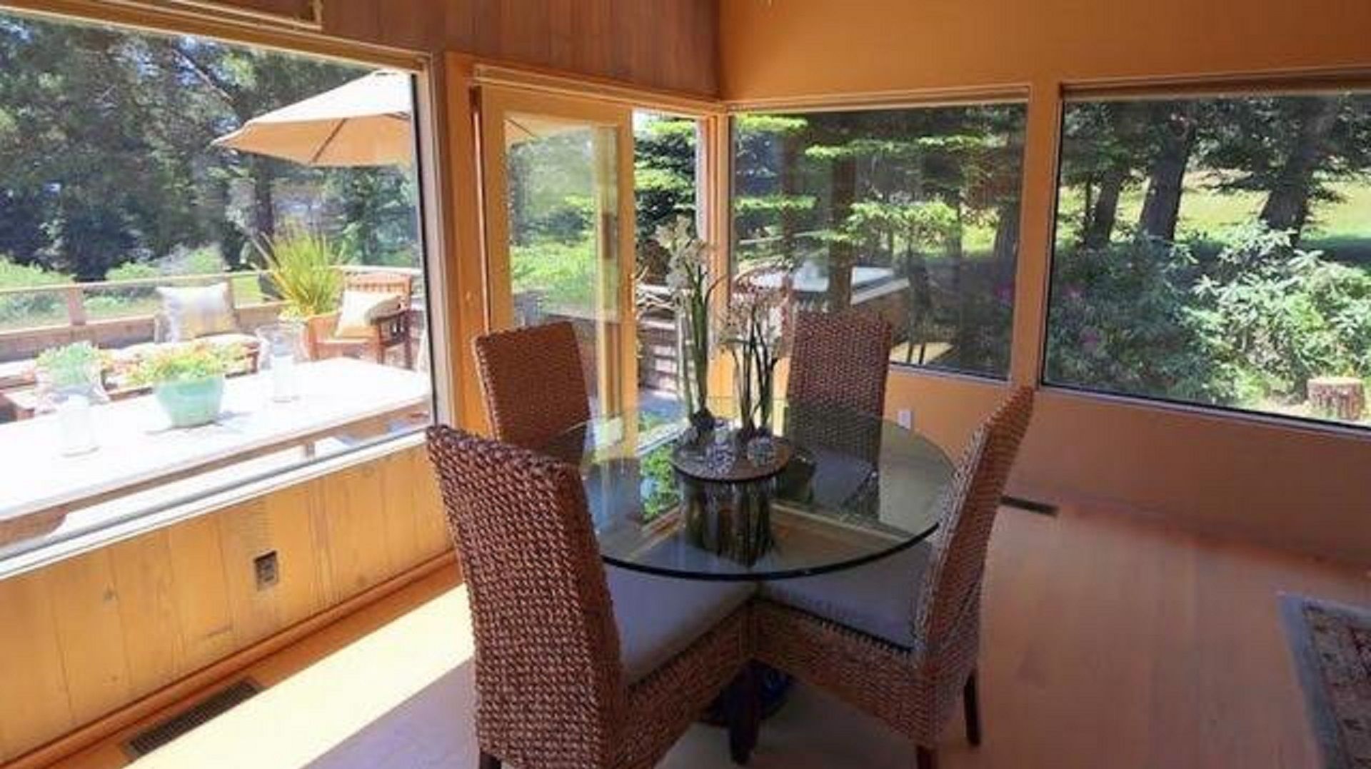 A glass-top dining table with wicker chairs, a view of a garden through large windows, and a wooden deck with outdoor furniture.