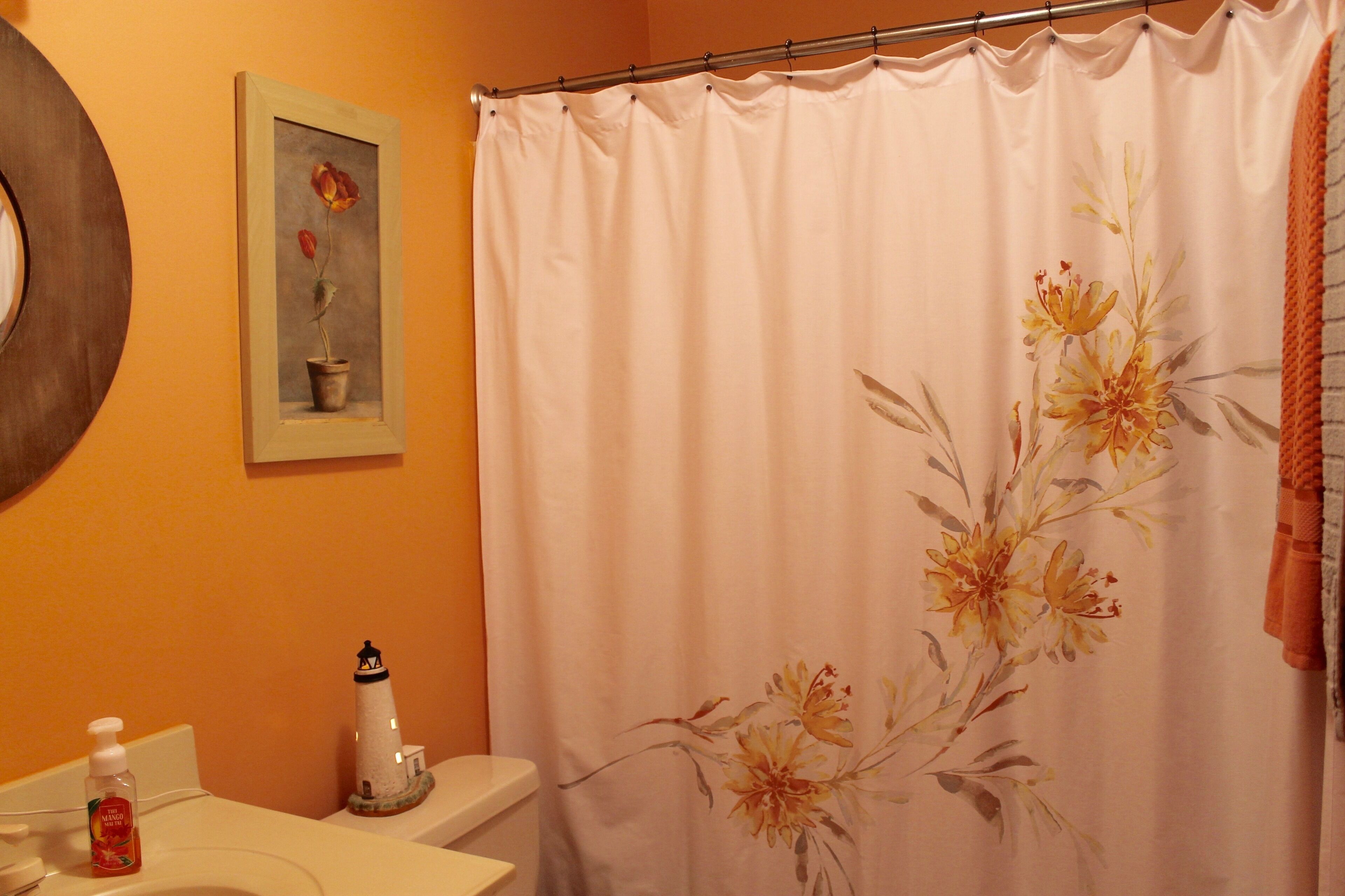 Une salle de bain avec un rideau de douche blanc, un tableau représentant une fleur, un flacon de savon liquide et une petite décoration en forme de phare.