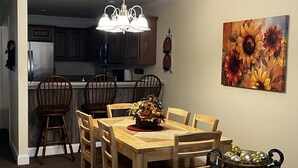 A dining area with a wooden table and chairs, a chandelier, a painting of sunflowers, and a kitchen in the background. - Luxury Condo In Town (Helen)