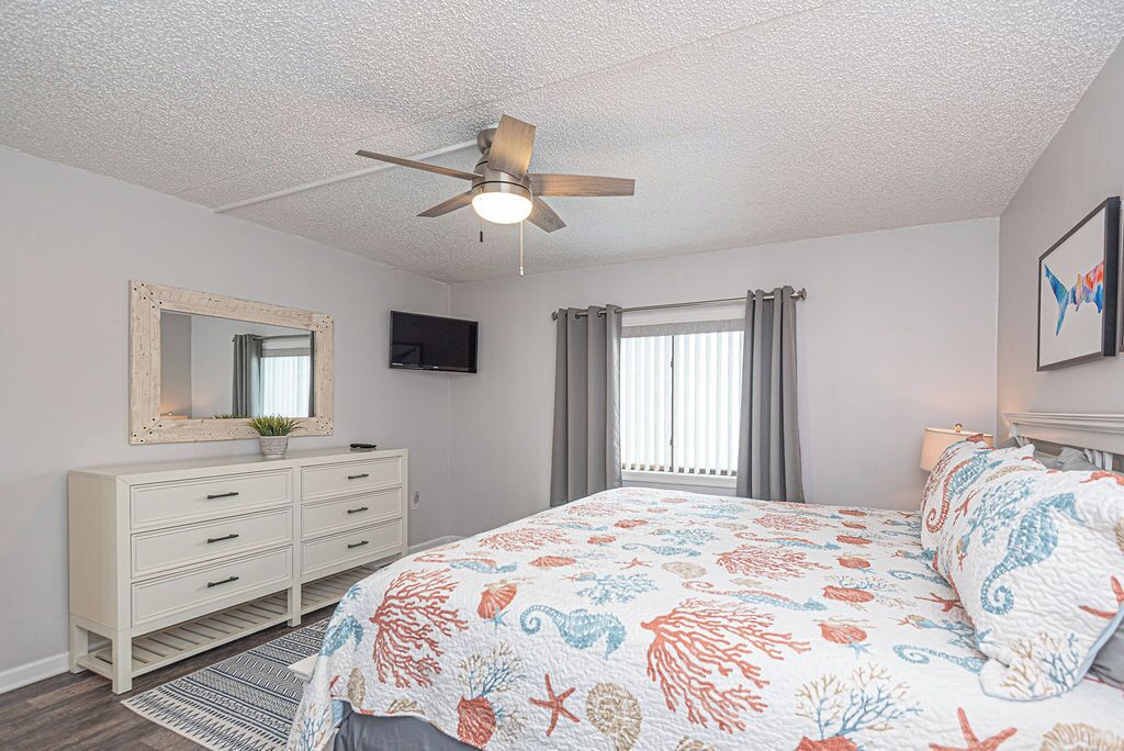 A bedroom with a bed, a ceiling fan, a dresser, a TV, and a window with curtains.