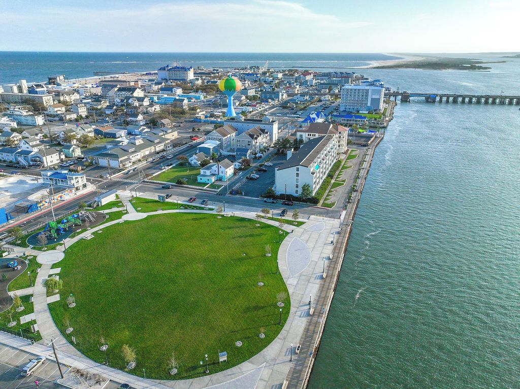 Vista aérea de un pueblo costero con un parque verde extenso, un paseo marítimo y diversos edificios, incluyendo una estructura azul destacada.