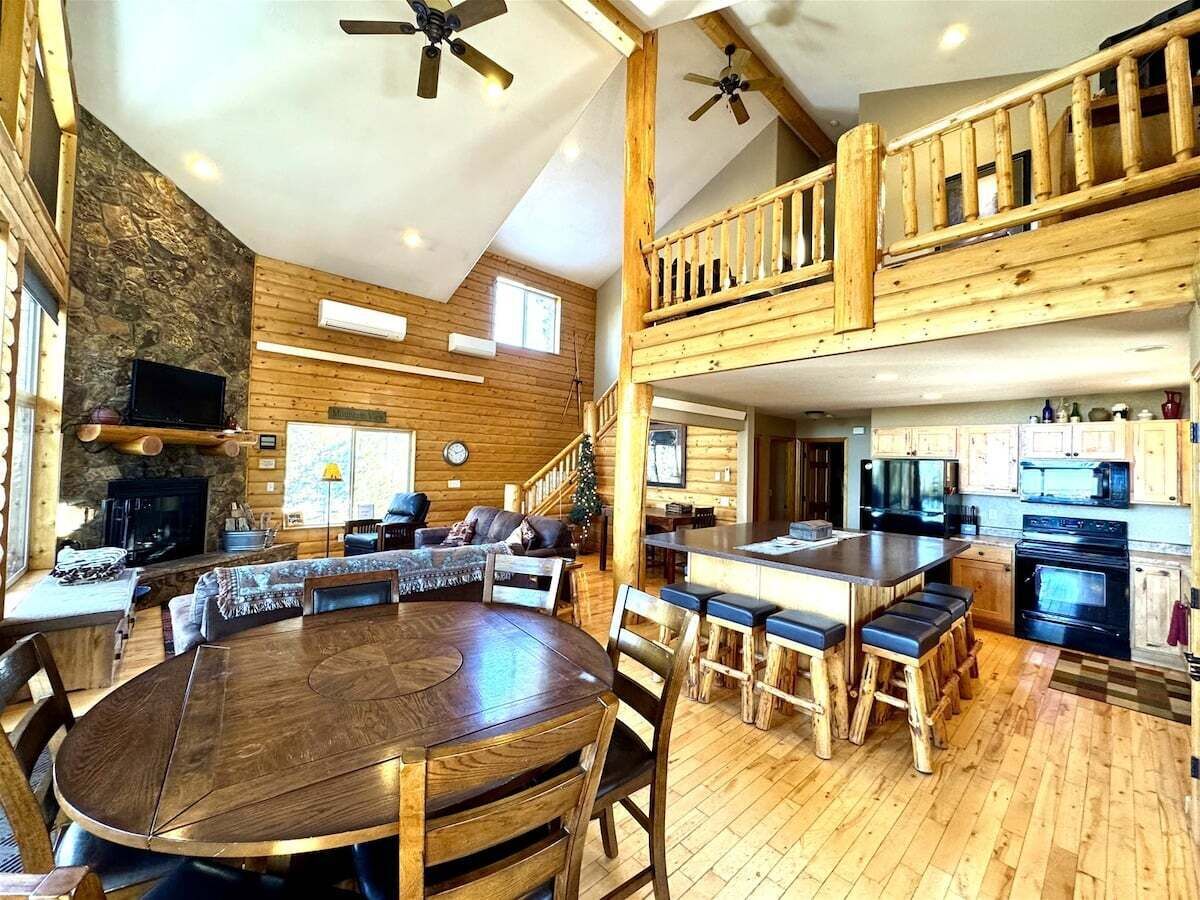 A spacious log cabin interior with a dining area, kitchen, and a loft bedroom.