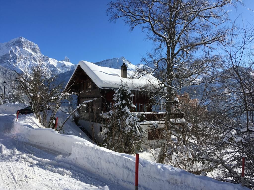 A wooden cabin with a snow-covered roof, surrounded by snow-covered trees and mountains in the background.