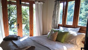 A wooden-framed bedroom with a bed, pillows, and a view of greenery outside.