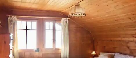 A cozy wooden cabin room with a bed, a window with curtains, and a hanging light fixture.