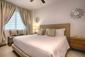 A bedroom with a large bed, a bedside table, a nightstand, a wall-mounted mirror, and a window with curtains. - Modern Mexican 1BR | 2 Pool, Gym & Balcony (Puerto Vallarta)