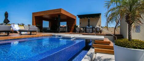 A swimming pool with a wooden deck, lounge chairs, and a covered seating area.