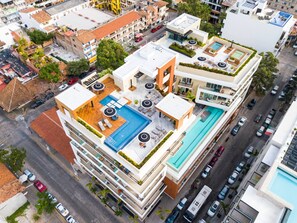 Un hotel de varios pisos con piscina en la azotea, mobiliario exterior y vista a la calle.