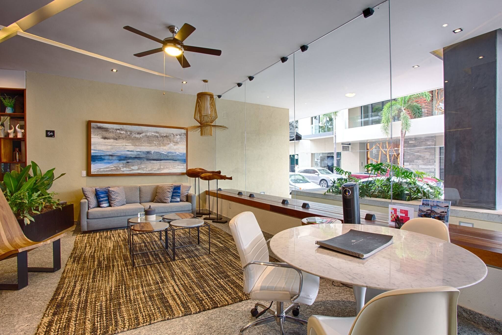Un salon moderne comprenant un canapé, une table basse et des fauteuils, un ventilateur de plafond et une grande fenêtre donnant sur la rue.