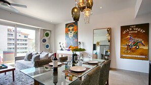 A modern living room with a glass dining table, grey sofa, and a large painting of a wild west scene. - Modern Mexican 1BR | 2 Pool, Gym & Balcony (Puerto Vallarta)