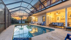 An indoor swimming pool with a glass ceiling, lounge chairs, and a patio area with a view of the sunset.