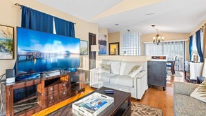 A modern living room with a large flat-screen TV, a white sofa, a dark wooden coffee table, and a patterned rug.