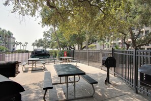 An outdoor patio area with picnic tables and a grill, surrounded by a metal fence and trees. - Direct Oceanfront Luxury, Miles of Ocean View, King bed, Walk-in Shower. (Hilton Head Island)