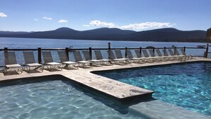 Una piscina cristalina con una cubierta de madera y sillones de descanso, con vistas a un lago y montañas al fondo.
