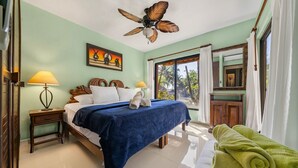 Quarto com cama, mesa de cabeceira, luminária, ventilador de teto, pintura, janela com cortinas e toalha verde.