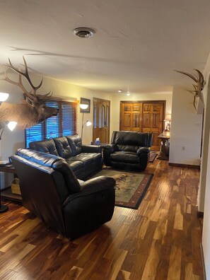 Ein Wohnzimmer mit einem großen Hirschgeweih an der Wand, Ledersofas, Holzböden und einer Holztür im Hintergrund.