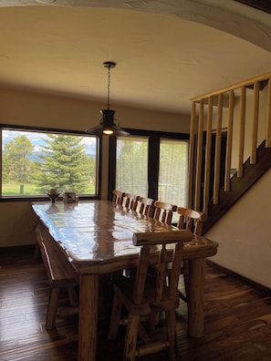 A wooden dining table with chairs, a hanging light fixture, a staircase, and large windows with a view of trees.