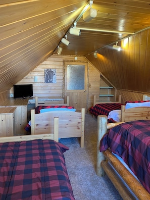 Um quarto de sótão acolhedor, com paredes de madeira, teto inclinado e uma televisão. Há três camas com roupa de cama xadrez e uma pequena janela.