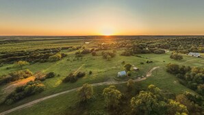 Une vue aérienne d’un paysage rural, avec des champs, des arbres et quelques bâtiments dispersés, prise au coucher du soleil.