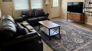 Un salon avec un canapé en cuir, une table basse en bois, un téléviseur posé sur un meuble en bois et un tapis à motifs.