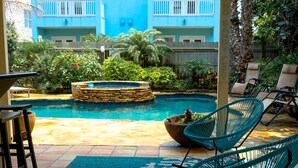 Zona de piscina con pared de piedra, sillas de descanso y vista a un edificio azul con balcones.
