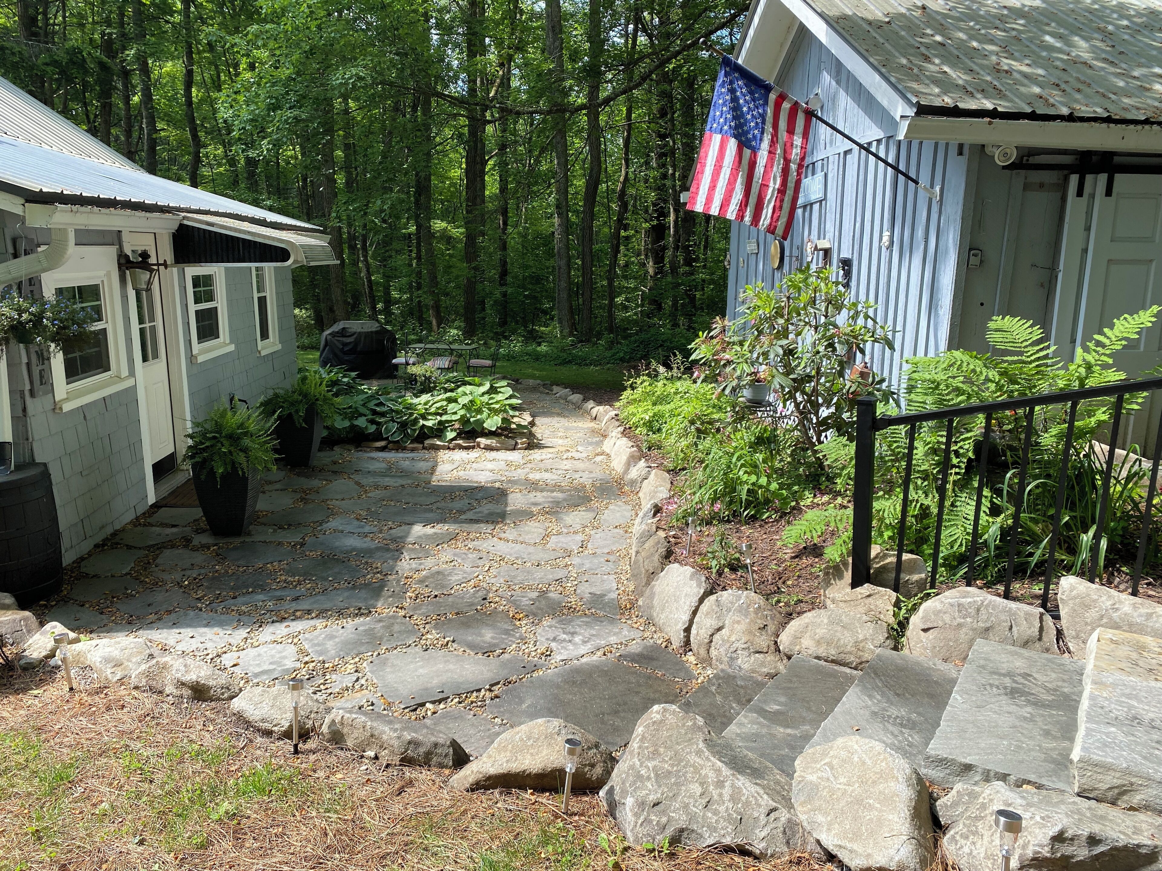 A stone pathway leading to a rustic cabin with an American flag, surrounded by lush greenery and a forested backdrop.