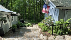 Un sentier de pierres mène à un chalet rustique orné d’un drapeau américain, le tout étant entouré d’une végétation luxuriante et bordé par une forêt dense.