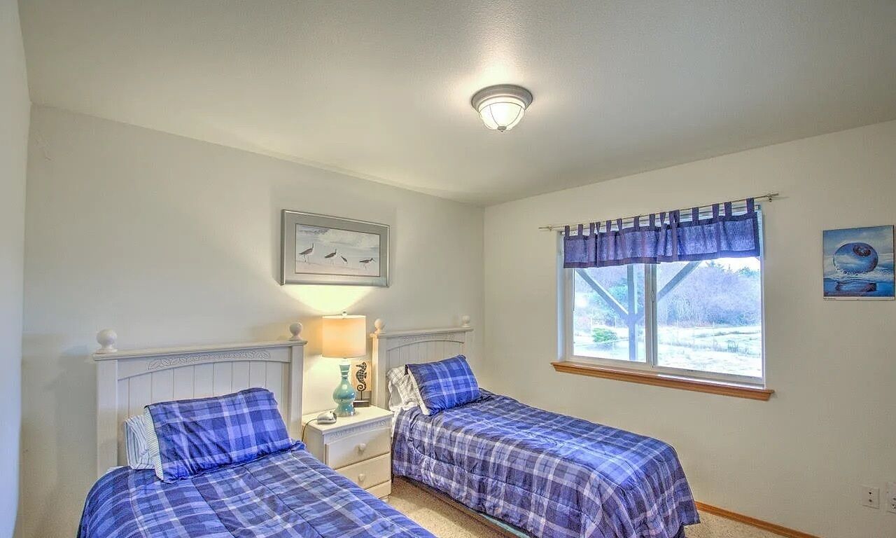 Une chambre avec deux lits, une table de chevet, une lampe, une fenêtre avec des rideaux et un tableau au mur.