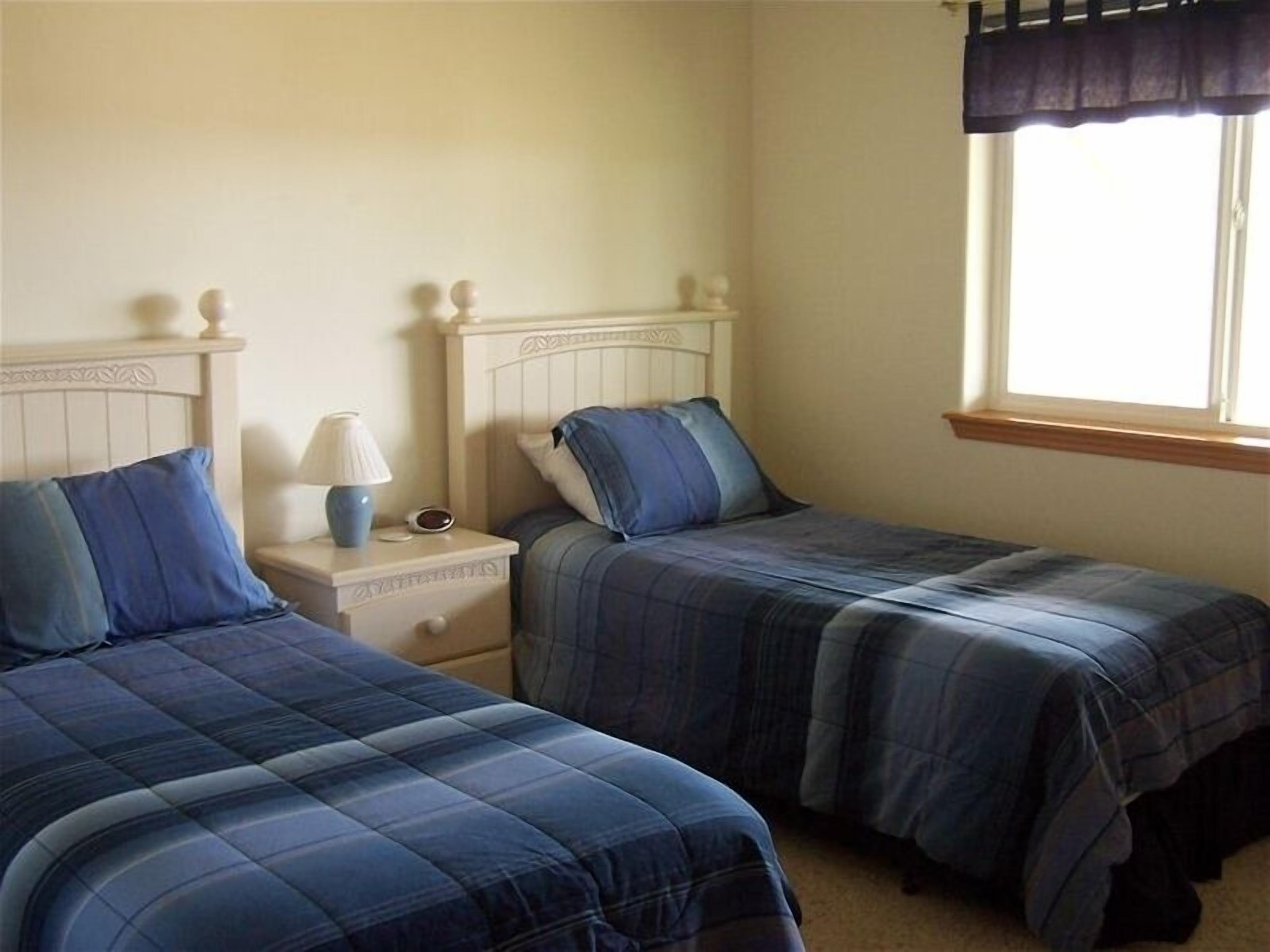 A bedroom with two single beds, a nightstand with a lamp, and a window with a dark valance.
