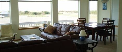 A living room with a brown leather couch, a dining table with chairs, and large windows offering a view of the outdoors.