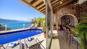 Un espace aménagé au bord de la piscine, avec vue sur la mer, une cheminée en pierre et des chaises longues.